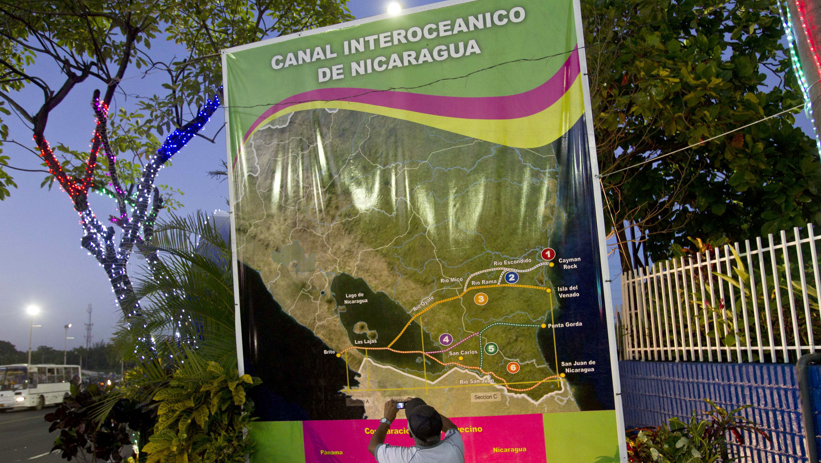 In this Dec. 4, 2013 photo, Oscar Torres, a 62-year-old retired construction worker takes photos of a banner showing a map of Nicaragua with possible routes of the Inter-Oceanic canal in Managua, Nicaragua. Six months after the Sandinista government granted a Chinese businessman a 100-year concession to build a vast canal across the country, Nicaraguans are confident the $40 billion canal will become reality, lifting their country from poverty to prosperity. (AP Photo/Esteban Felix)