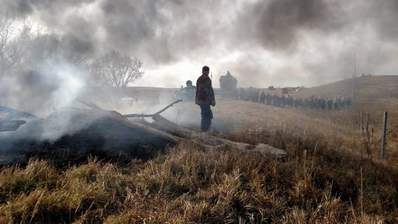 A water protector stares down police along County Road 134, in Mandan, North Dakota, north of Red Warrior Camp. Law enforcement wear riot gear as they prepare to remove the frontline camp. Oct. 27, 2016 (Derrick Broze for MintPress) 