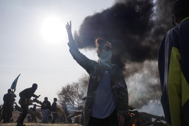 Gabriella Scarlett, a water protector from Canada, signals for peace as a fire barricade burns off County Road 134. Behind her, water protectors establish a fire barricade to hold police back from the site of construction of the Dakota Access pipeline. Oct. 27, 2016 (Derrick Broze for MintPress) 