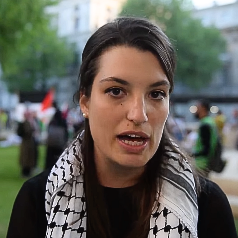 Deborah Fiorin speaking at a protest, wearing a black top and Palestinian keffiyeh
